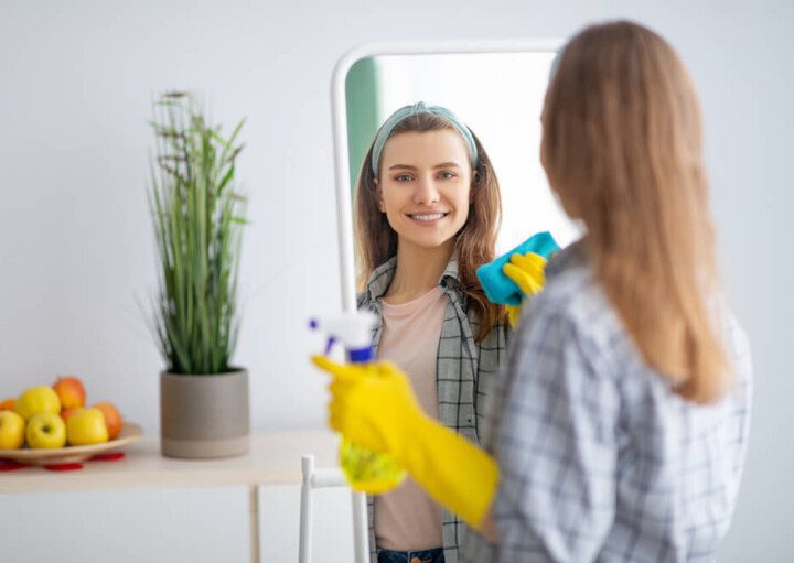 Foto que ilustra matéria com dicas sobre como limpar espelho mostram uma mulher de costas, posicionada de frente para um espelho. Ela aparece sorrindo no reflexo, com um luvas amarelas, com um pano verde esfregando o espelho em uma das mãos e um borrifador na outra.