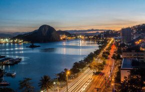 Foto que ilustra matéria sobre o custo de vida em Vitória (ES) mostra uma visão do alto da cidade no cair da tarde, em que aparece uma parte da água da baía à esquerda da tela, com um morro ao fundo, ao centro uma avenida e mais à esquerda prédios residenciais.