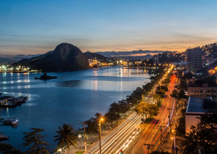 Foto que ilustra matéria sobre o custo de vida em Vitória (ES) mostra uma visão do alto da cidade no cair da tarde, em que aparece uma parte da água da baía à esquerda da tela, com um morro ao fundo, ao centro uma avenida e mais à esquerda prédios residenciais.