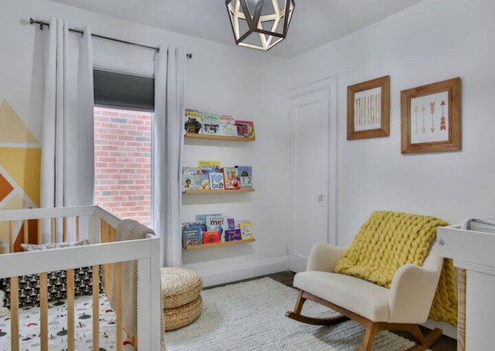 quarto de bebê decorado em tons de branco e mostarda. Há um berço, uma janela coberta por cortinas, uma poltrona e uma cômoda.