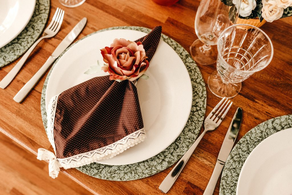 Mesa posta de jantar com talheres, taças, sousplat, prato, guardanapo em poá e porta-guardanapo em formato de flor.