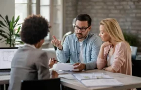 Imagem de um homem e uma mulher sentados em uma mesa com um profissional, discutindo documentos relacionados à Lei do inquilinato. O homem gesticula enquanto fala, a mulher ouve atentamente, e papéis e uma planta baixa estão espalhados sobre a mesa.