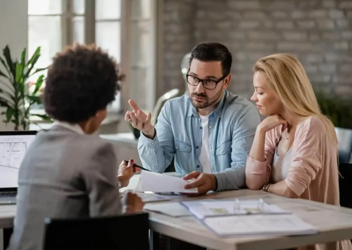 Imagem de um homem e uma mulher sentados em uma mesa com um profissional, discutindo documentos relacionados à Lei do inquilinato. O homem gesticula enquanto fala, a mulher ouve atentamente, e papéis e uma planta baixa estão espalhados sobre a mesa.