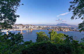 Foto que ilustra matéria sobre Morar em Vitória (ES) mostra uma visão do alto da cidade, em que uma vegetação aparece em primeiro plano, com uma parte da baía da cidade no meio, uma de suas pontes mais à esquerda, além de prédios e montanhas mais ao fundo.