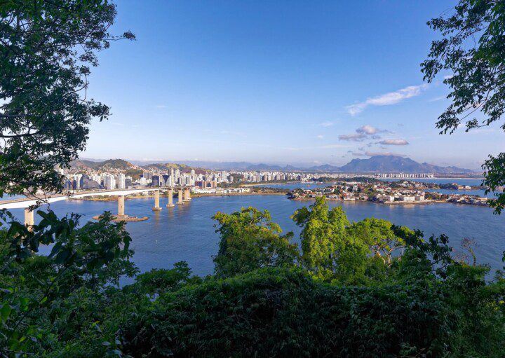 Foto que ilustra matéria sobre Morar em Vitória (ES) mostra uma visão do alto da cidade, em que uma vegetação aparece em primeiro plano, com uma parte da baía da cidade no meio, uma de suas pontes mais à esquerda, além de prédios e montanhas mais ao fundo.