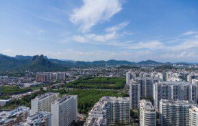 Foto que ilustra matéria sobre a Zona Oeste do RJ mostra uma vista aérea de Jacarepaguá, com grandes condomínios de prédios residenciais em primeiro plano e montanhas ao fundo.