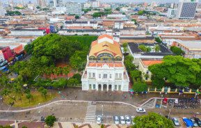 Foto que ilustra matéria sobre o que fazer em Fortaleza mostra o Theatro José de Alencar visto de cima. A construção branca, com um topo colorido onde fica o letreiro, é cercada por árvores e tem casas e prédios da cidade ao fundo.