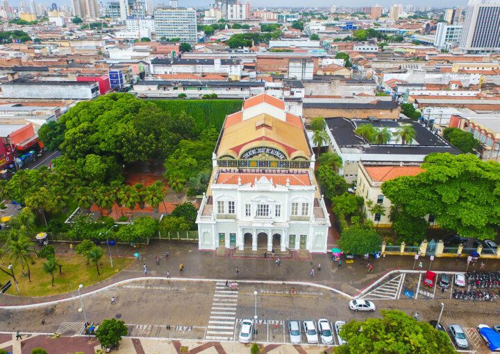 Foto que ilustra matéria sobre o que fazer em Fortaleza mostra o Theatro José de Alencar visto de cima. A construção branca, com um topo colorido onde fica o letreiro, é cercada por árvores e tem casas e prédios da cidade ao fundo.