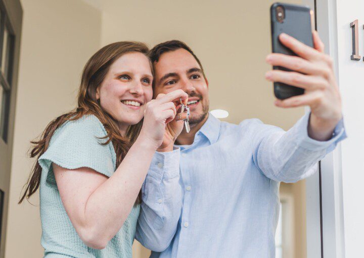 Foto que ilustra matéria sobre como montar uma carteira de investimentos para comprar um imóvel mostra um jovem casal tirando uma foto com o celular mostrando as chaves da casa nova