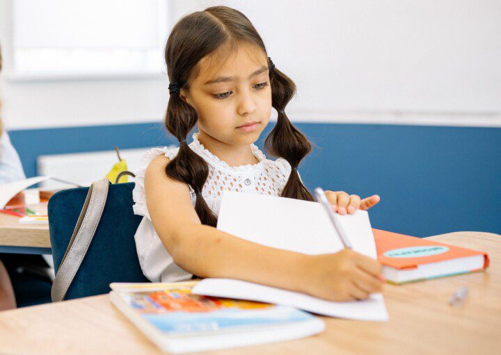 Foto que ilustra matéria sobre Escolas em Fortaleza mostra uma menina sentada com um caderno aberto à sua frente e escrevendo com um lápis em uma sala de aula.