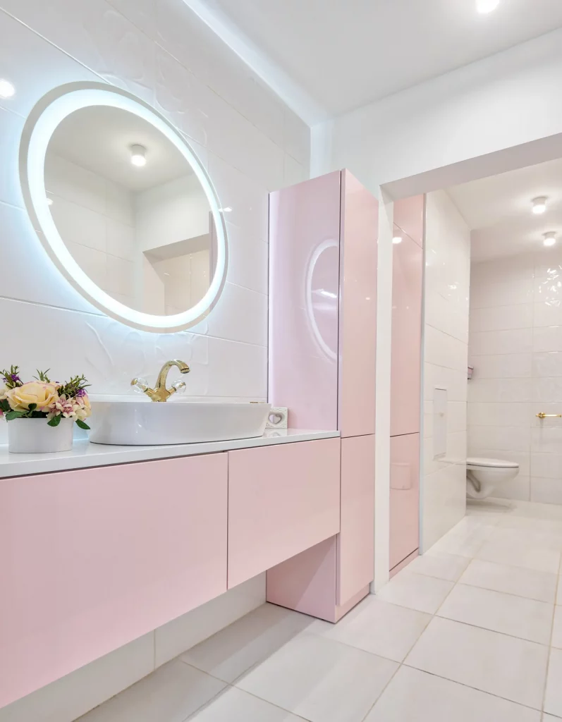 Imagem de um lavabo moderno com tons de rosa, uma pia com gabinete e um espelho circular com luz led no seu contorno.