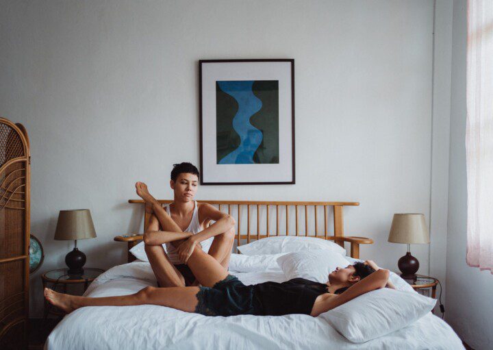 A imagem contém duas mulheres deitadas em uma cama no quarto de casal. Uma está de camiseta regata branca e a outra preta. No quarto há um quadro na parede ao fundo da cama, uma cabeceira de madeira, uma mesinha de cabeceira de cada lado da cama com abajur.