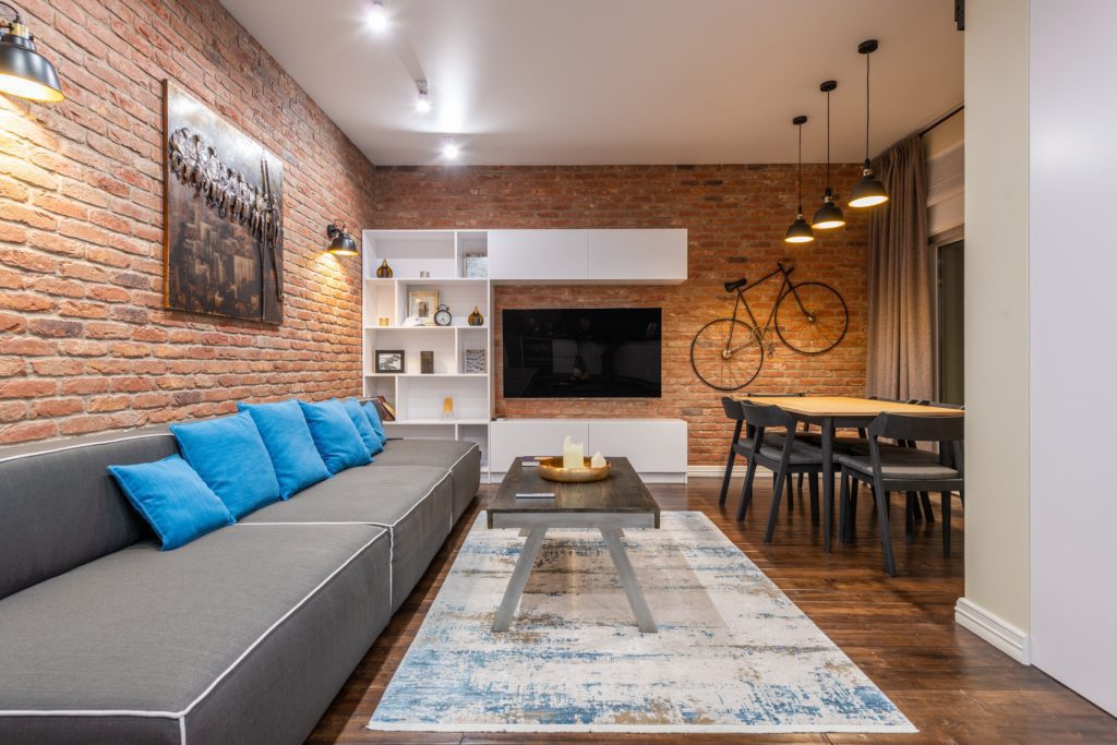 Imagem de uma decoração de sala grande com um tapete no centro, um grande sofá cinza com almofadas azuis no canto esquerdo, uma mesa no centro e as paredes de tijolinho. A sala ainda conta com uma estante branca no fundo com a TV pendurada na parede e uma bicicleta também pendurada na parede para compor a decoração de sala grande.