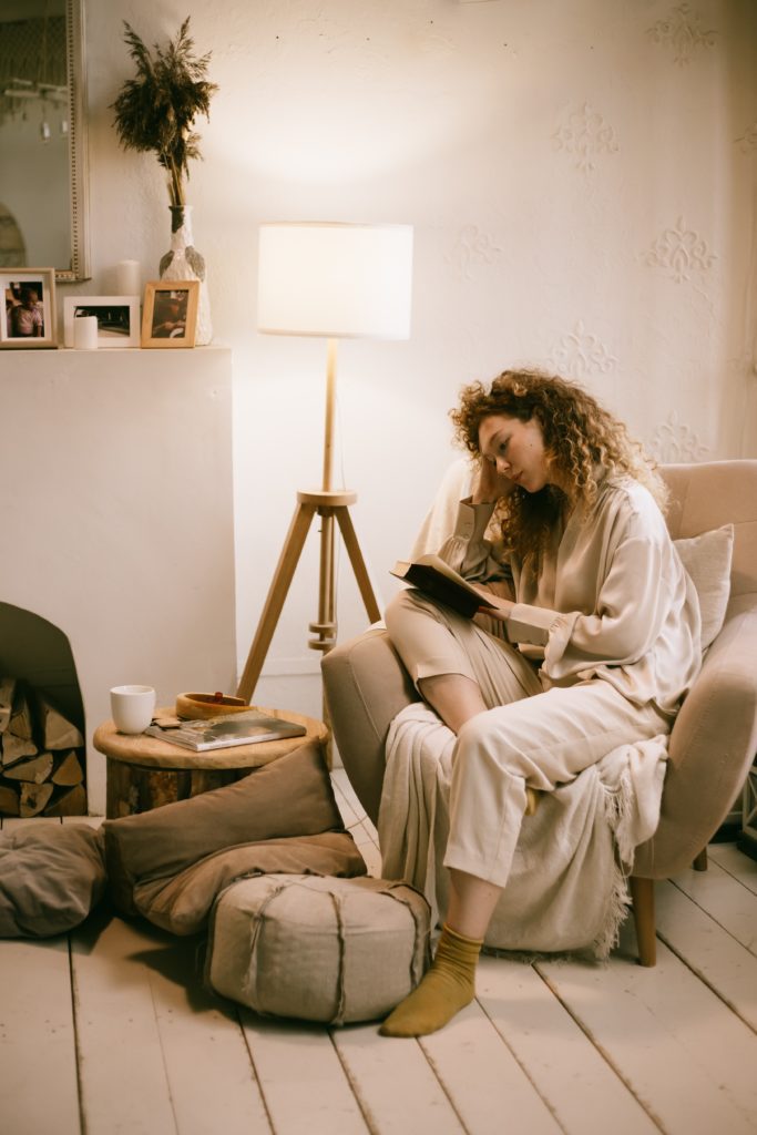  Imagem de um espaço para a leitura com uma moça sentada em uma poltrona com roupas cinzas e meia, uma mesinha no canto e uma luminária de chão ao fundo, e ainda contém um espaço com fotos na parede.