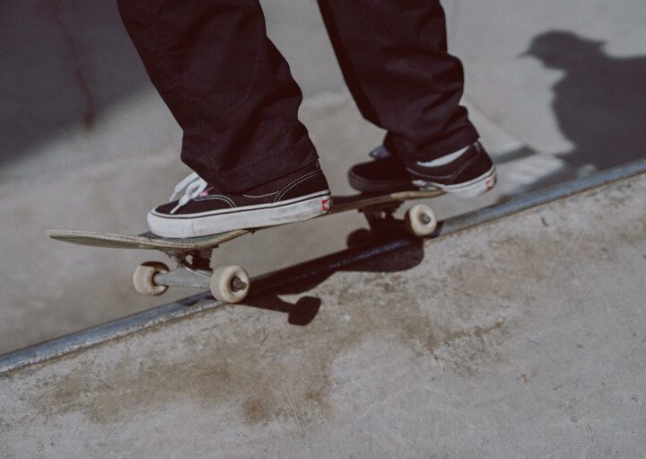 Foto que ilustra matéria sobre Pista de skate em Santos mostra um detalhe de um skate sendo equilibrado na borda de uma pista, com dois pés de um skatista sobre ele. O skatista usa calças compridas pretas e um tênis preto com parte do solado branco