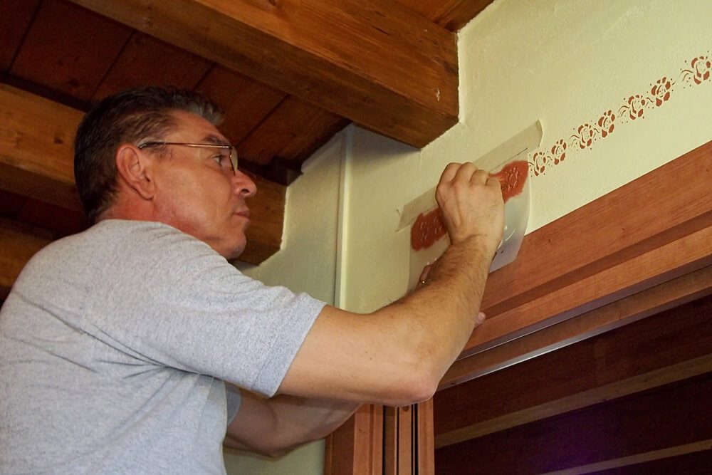 Foto que ilustra matéria sobre como fazer stencil mostra um homem pintando uma parede com pequenas flores usando um molde e um pincel fino.