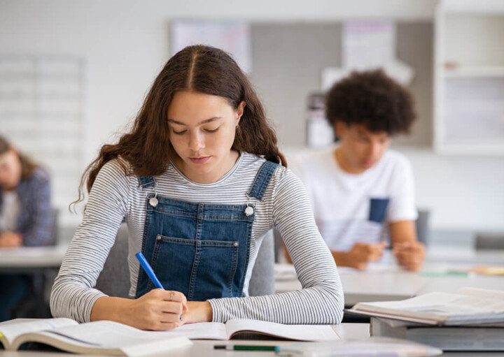 Foto que ilustra matéria sobre escolas em Santos mostra uma adolescentes de cabelos compridos pretos e cacheados, usando um macacão jeans com uma blusa cinza de mangas compridas por baixo, sentada a uma carteira de escola escrevendo em um caderno.