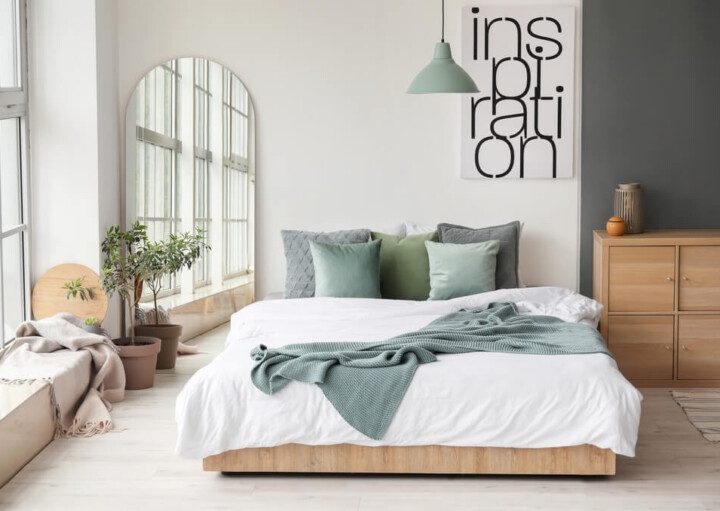 Foto que ilustra matéria sobre parede de quarto decorada mostra um quarto com uma grande cama ao centro, com algumas almofadas em cima. À esquerda, ao lado da cama, há um grande espelho. E acima da cama, na parede, no canto superior direito, há um quadro com a palavra “inspiration”.