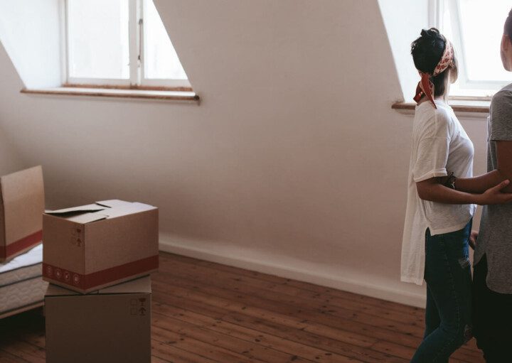 Foto que ilustra matéria sobre o Censo QuintoAndar com foco em desejos de mudança de casa mostra um casal abraçado em um quarto, de costas, olhando por uma janela. E ao lado esquerdo uma cama com algumas caixas de papelão em cima em no chão à frente dela