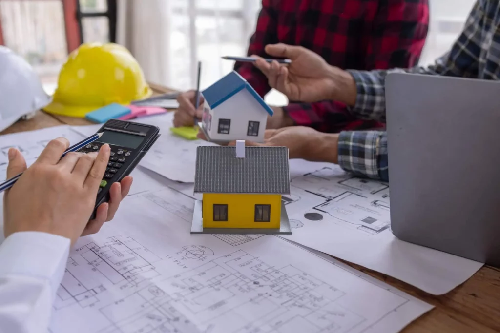 Imagem de pessoas ao redor de uma mesa onde estão algumas casas em tamanho miniatura e plantas arquitetônicas. Também estão visíveis uma calculadora, um notebook, um tablet e capacetes de segurança amarelos