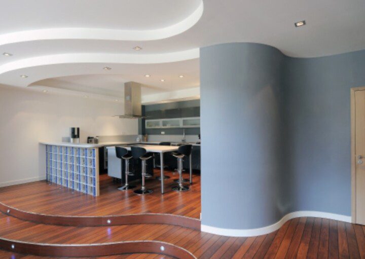 Foto que ilustra matéria sobre decoração de paredes curvas mostra a entrada de uma sala com chão de madeira e uma parede curva pintada de azul. Ao lado dela, uma cozinha americana, com uma mesa e banquetas.