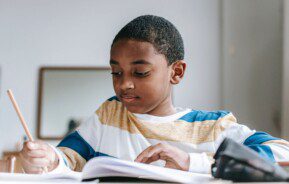 Foto que ilustra matéria sobre escolas em Santo André mostra um menino negro sentado à uma mesa enquanto escreve de lápis em um caderno.