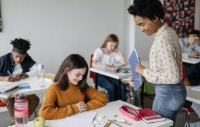 Foto que ilustra matéria sobre Escolas em Goiânia mostra uma menina sentada em uma carteira de escola enquanto é atendida por uma professora que está de pé a sua frente.