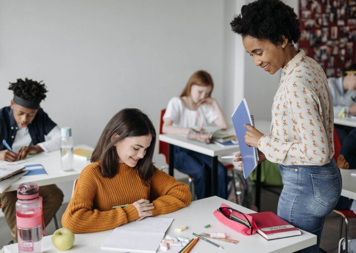 Foto que ilustra matéria sobre Escolas em Goiânia mostra uma menina sentada em uma carteira de escola enquanto é atendida por uma professora que está de pé a sua frente.