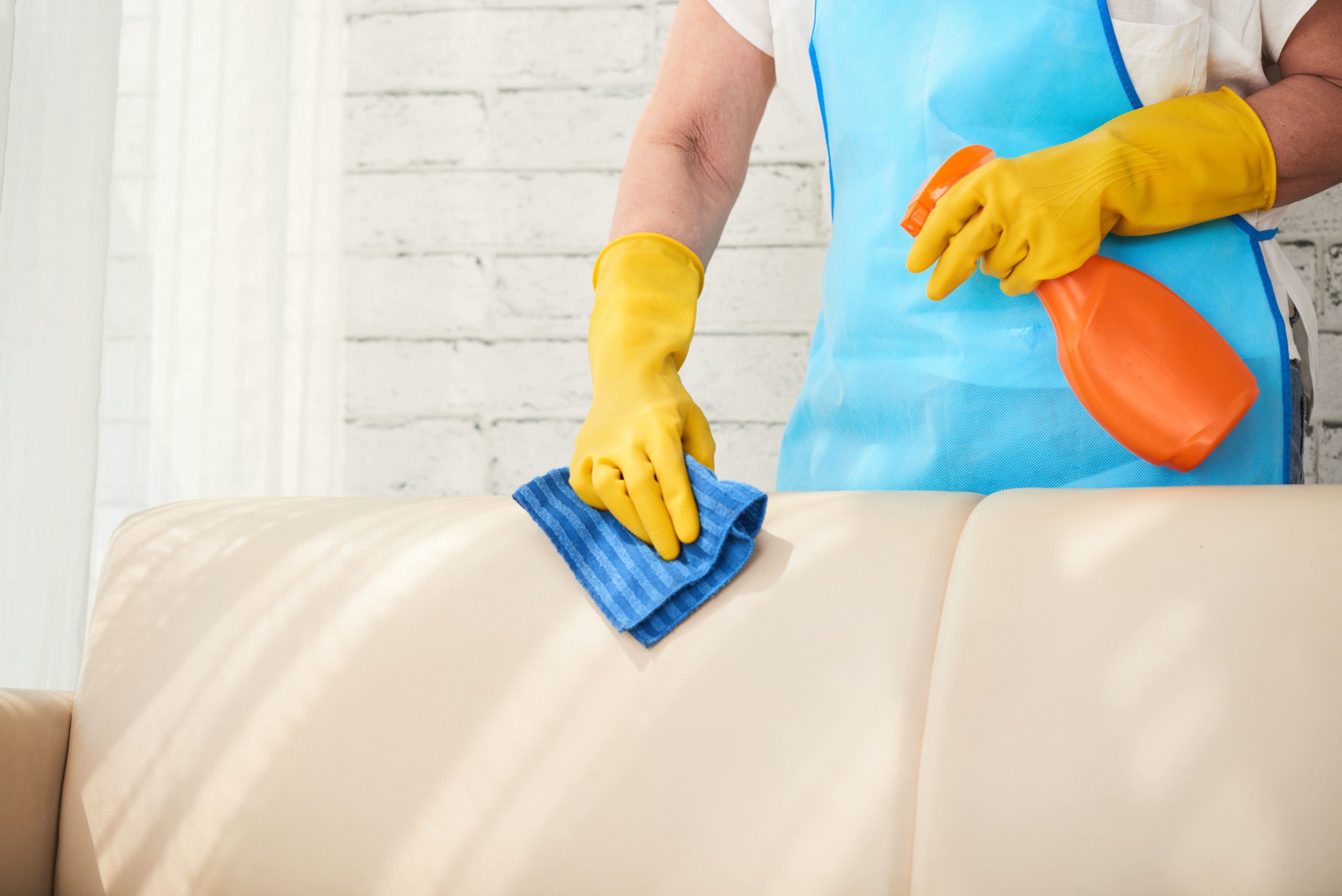 Limpeza e higienização de sofá com borrifador e pano umido sendo feita com luvas de proteção.