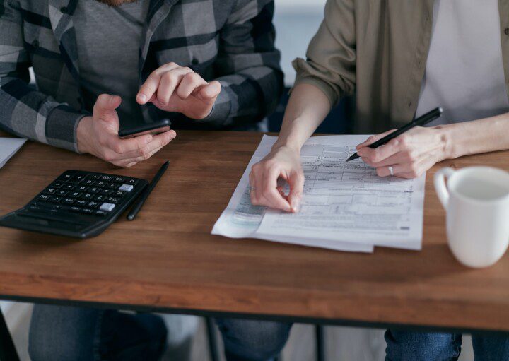 Foto que ilustra matéria sobre Tabela Price ou SAC, qual é melhor para o financiamento imobiliário, mostra duas pessoas fazendo contas com calculadora, celular e papel em uma mesa de madeira.