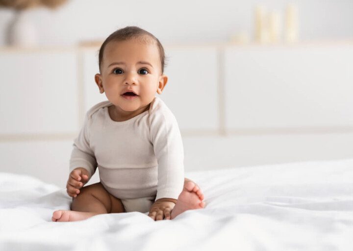 Foto que ilustra matéria sobre quarto de bebê simples mostra um bebê de poucos meses sentado em cima de uma cama olhando para a câmera. Ao fundo, desfocados, aparecem alguns móveis brancos.