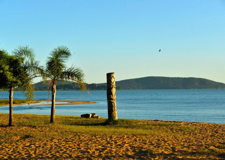 Foto que ilustra matéria sobre as praias de Porto Alegre mostra um trecho da praia de Ipanema, onde aparecem uma faixa de areia em primeiro plano, alguns coqueiros à esquerda, o espelho d’água no meio da imagem e pequenos morros ao fundo.