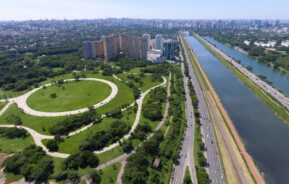 Foto que ilustra matéria sobre a Zona Oeste de SP mostra uma panorâmica da cidade, feita do alto, com destaque para o Parque Villa-Lobos, uma área verde que aparece no canto inferior esquerdo, a Marginal e o Rio Pinheiros em todo o canto direito e prédios e casas ao fundo no canto superior esquerdo da imagem.