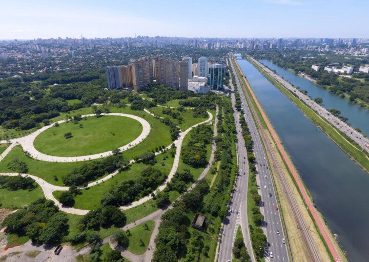 Foto que ilustra matéria sobre a Zona Oeste de SP mostra uma panorâmica da cidade, feita do alto, com destaque para o Parque Villa-Lobos, uma área verde que aparece no canto inferior esquerdo, a Marginal e o Rio Pinheiros em todo o canto direito e prédios e casas ao fundo no canto superior esquerdo da imagem.