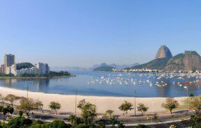 Foto que ilustra matéria sobre a Zona Sul do RJ mostra a Enseada de Botafogo, com carros passando na pista logo abaixo da imagem, uma faixa de areia, o espelho d’água com vários barcos e o Pão de Açúcar ao fundo.