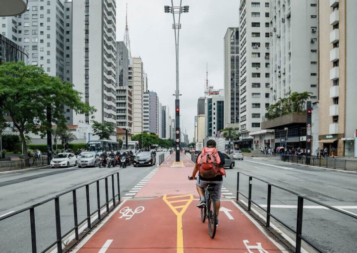 Foto que ilustra matéria sobre ciclovia em SP mostra a faixa vermelha destinada a ciclistas no canteiro central da Avenida Paulita. Aos lados estão as faixas de rolamento para carros e os prédios. E a imagem mostra um ciclista de costas, pedalando com uma mochila vermelha.