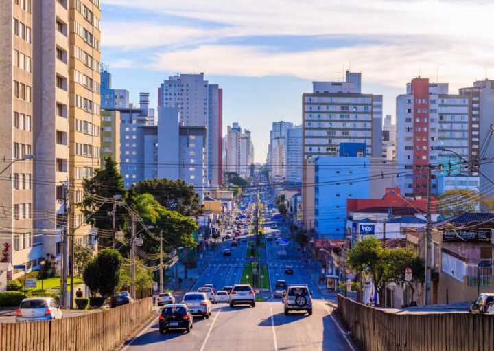 Foto que ilustra matéria sobre custo de vida em Curitiba mostra uma grande avenida do centro da cidade, com carros passando na via ao centro e altos prédios em volta.