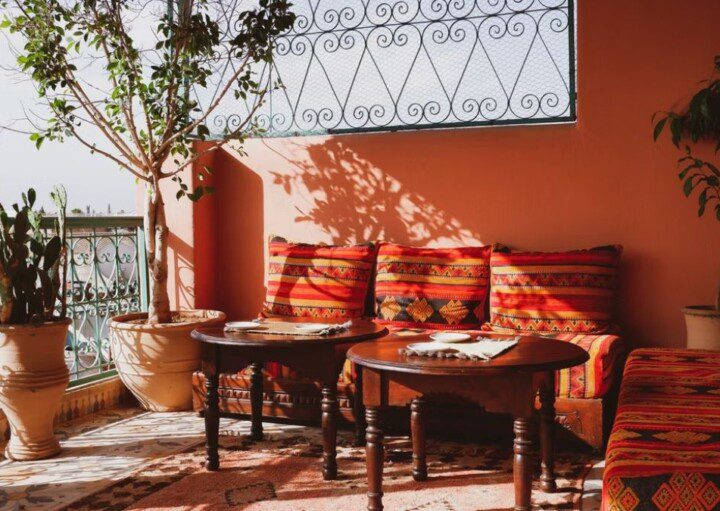 decoração de varanda com tons laranjas, sofás com almofadas listradas, mesas de centro, e a iluminação batendo em parte do ambiente. Há plantas decorando as beiras.