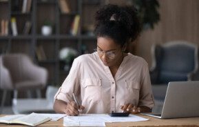 Foto que ilustra matéria sobre juros de mora mostra uma mulher negra, sentada a uma mesa, com um notebook aberto de lado, fazendo contas em uma calculadora e escrevendo em um papel.