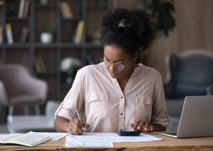 Foto que ilustra matéria sobre juros de mora mostra uma mulher negra, sentada a uma mesa, com um notebook aberto de lado, fazendo contas em uma calculadora e escrevendo em um papel.