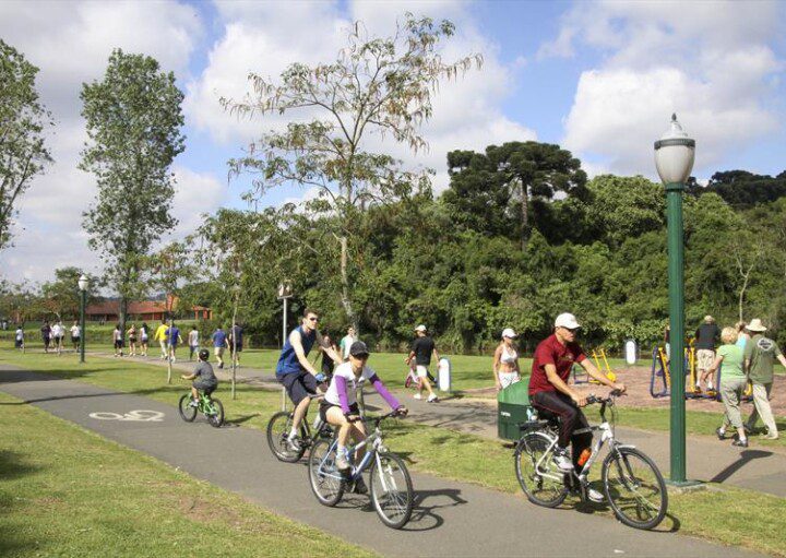 Foto que ilustra matéria sobre ciclovias em Curitiba mostra uma pista de asfalto repleta de ciclistas em meio a um parque arborizado.