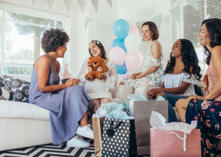 Foto que ilustra matéria sobre decoração para chá de bebê mostra cinco mulheres sentadas em dois sofás. Elas estão sorridentes e cercadas de presentes. A segunda mulher da esquerda para direita está vestida de branco e grávida e segura um ursinho de pelúcia. No centro da imagem, alguns balões nas cores azul, branco e rosa decoram o ambiente.