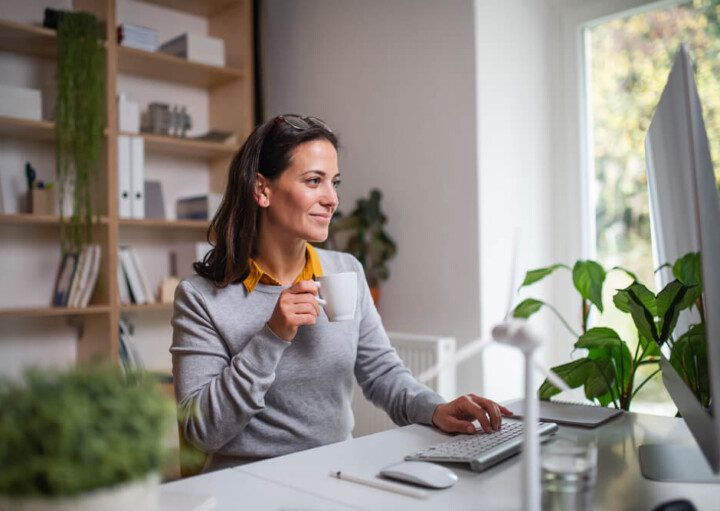 Foto que ilustra matéria sobre decoração para escritório mostra uma mulher sentada a uma mesa em casa, ao lado de uma grande janela. Ela segura uma xícara de café. Em cima da mesa, há monitor, teclado e mouse e alguns vasinhos de plantas. E atrás dela uma estante com livros, arquivos e plantas de decoração.