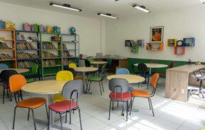 Foto que ilustra matéria sobre escolas em Osasco mostra uma sala de estudos do Colégio Cunha Carvalho, com diversas mesas redondas de madeira cercadas por cadeiras com estofados de diferentes cores. Ao fundo, há estantes com diversos livros.