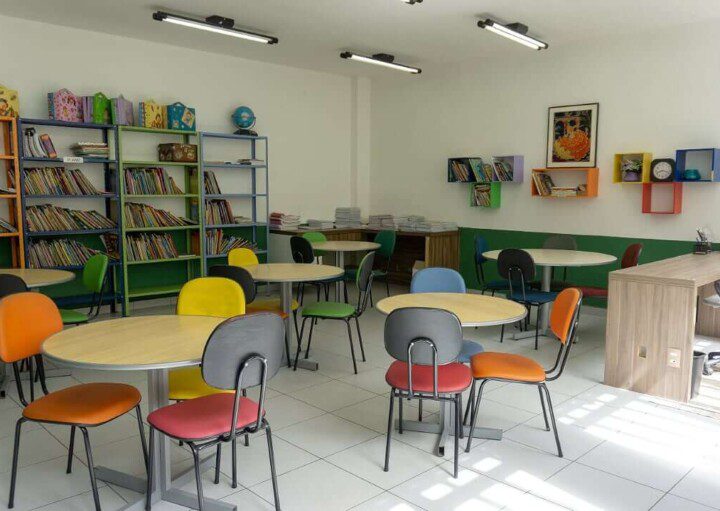 Foto que ilustra matéria sobre escolas em Osasco mostra uma sala de estudos do Colégio Cunha Carvalho, com diversas mesas redondas de madeira cercadas por cadeiras com estofados de diferentes cores. Ao fundo, há estantes com diversos livros.