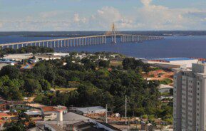 Foto que ilustra matéria sobre os melhores bairros de Manaus mostra ao fundo um grande espelho d’água formado pelo Rio Negro com uma ponte cruzando a image de uma ponta à outra. Do centro da foto para baixo, à margem do rio, há uma faixa de terra com árvores, algumas casas e um prédio.