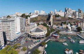 Foto que ilustra matéria sobre as melhores cidades do Nordeste para morar mostra uma vista panorâmica de Salvador, na Bahia, com detalhe para o Mercado Modelo, no centro da imagem, com o Elevador Lacerda um pouco atrás. No canto inferior direito, observa-se uma parte da baía, com barcos. À esquerda do mercado, estão alguns prédios, assim como no fundo, na parte alta da cidade. Mais ao fundo, o céu está azul.