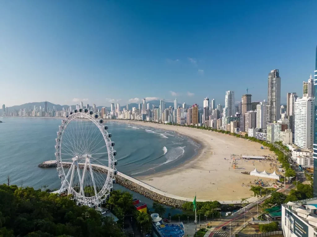 Imagem da vista aérea do litoral de Balneário Camboriú com uma roda gigante próximo ao mar para ilustrar matéria sobre quais são as melhores cidades de Santa Catarina para morar