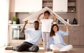 Imagem de uma família formada por um homem, uma mulher e uma criança sentados no chão de uma cozinha com um papelão na cabeça que simboliza o teto de uma casa para ilustrar matéria sobre caução de aluguel