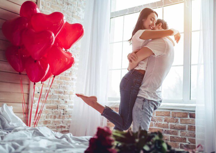 Casal de namorados se abraçando. O quarto está com decoração para o dia dos namorados.